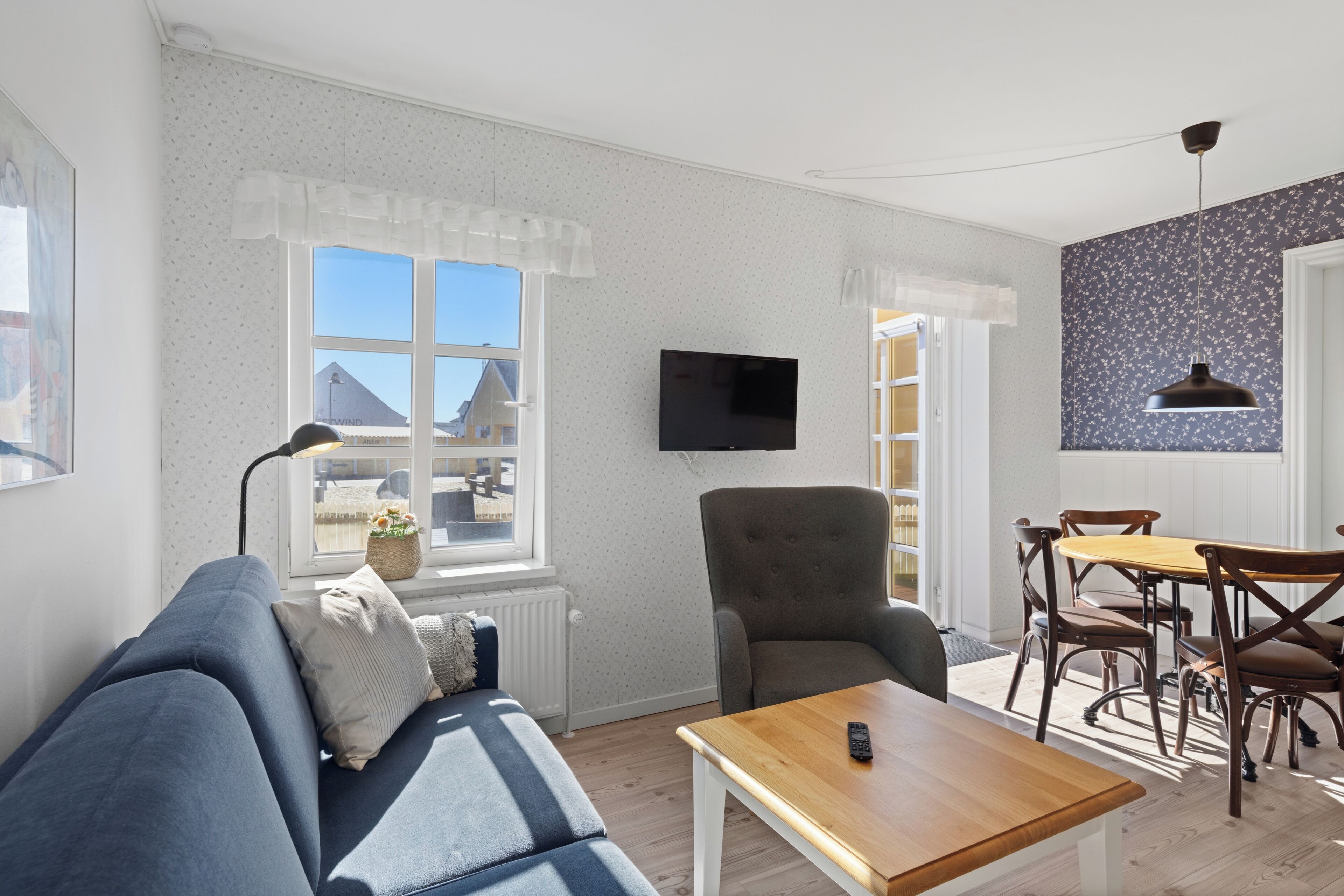 A living room with a sofa, armchair, and coffee table. There is a television mounted on the wall and a dining area with chairs and a table.
