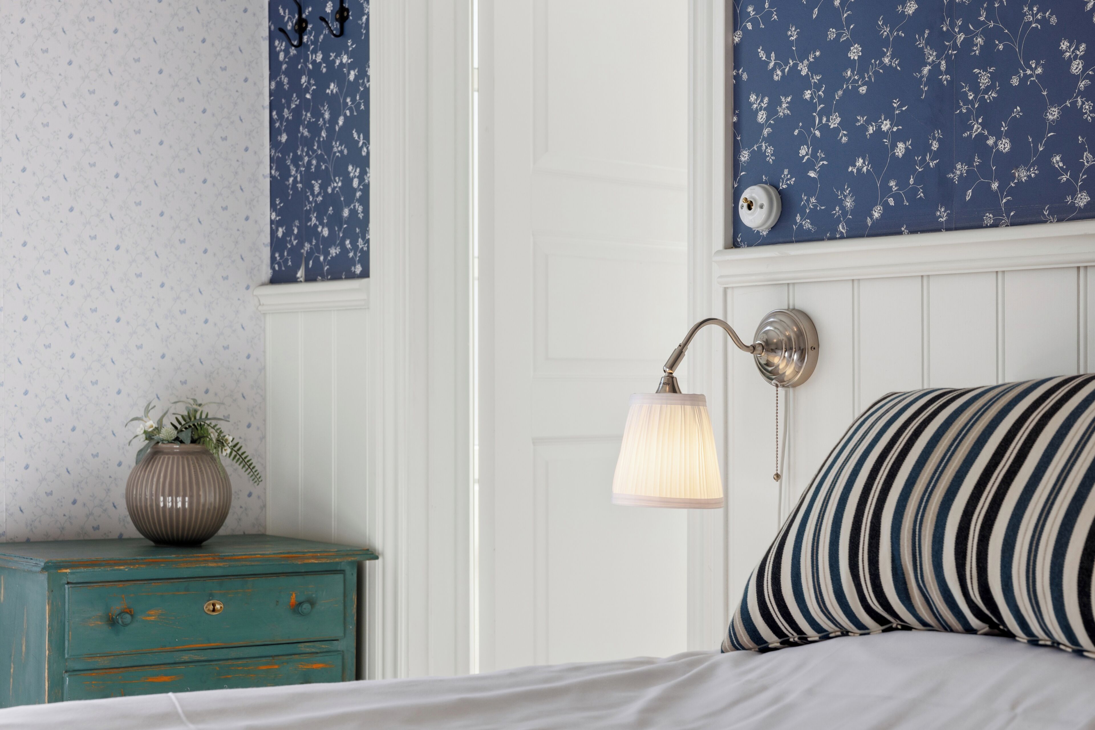 A bedroom with a bed, a nightstand with a vase, a wall-mounted lamp, and a door.