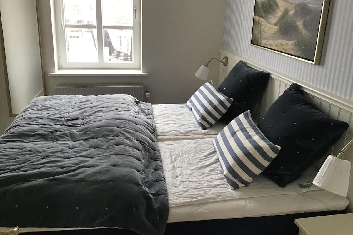 Une chambre à coucher avec un lit, deux oreillers, une table de chevet, une lampe, une fenêtre et un tableau au mur.