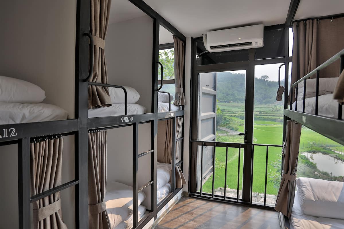 A dormitory room with bunk beds, an air conditioning unit, and a window overlooking a green landscape.