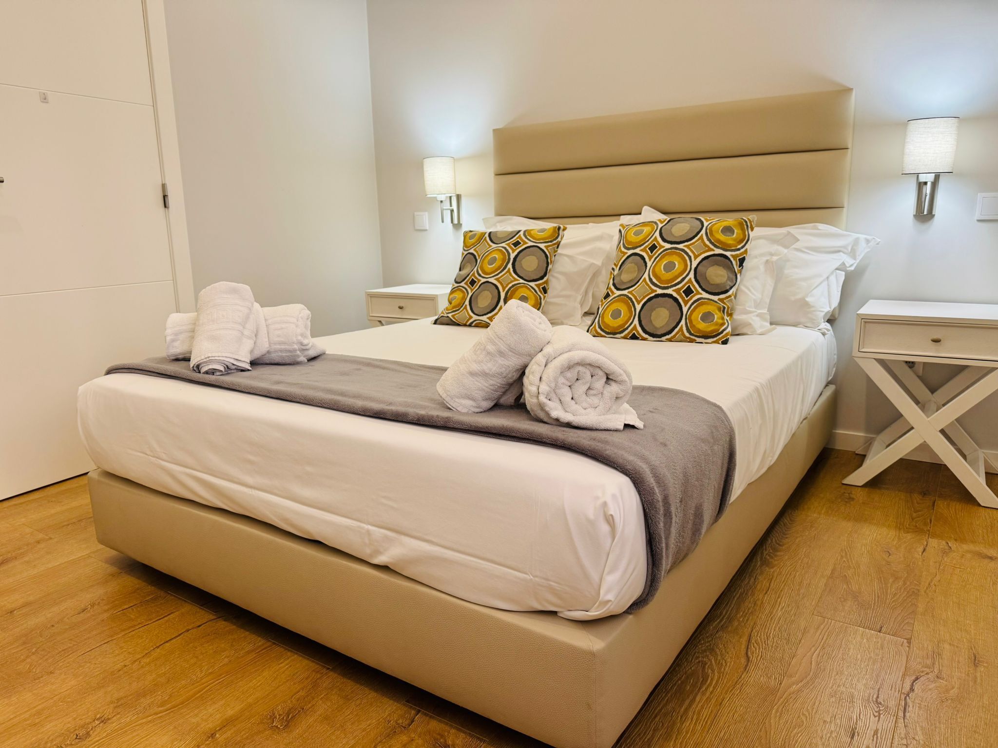 Una cama bien tendida con sábanas blancas, una manta gris y toallas enrolladas. Hay dos mesitas de noche con lámparas, y el suelo es de madera.