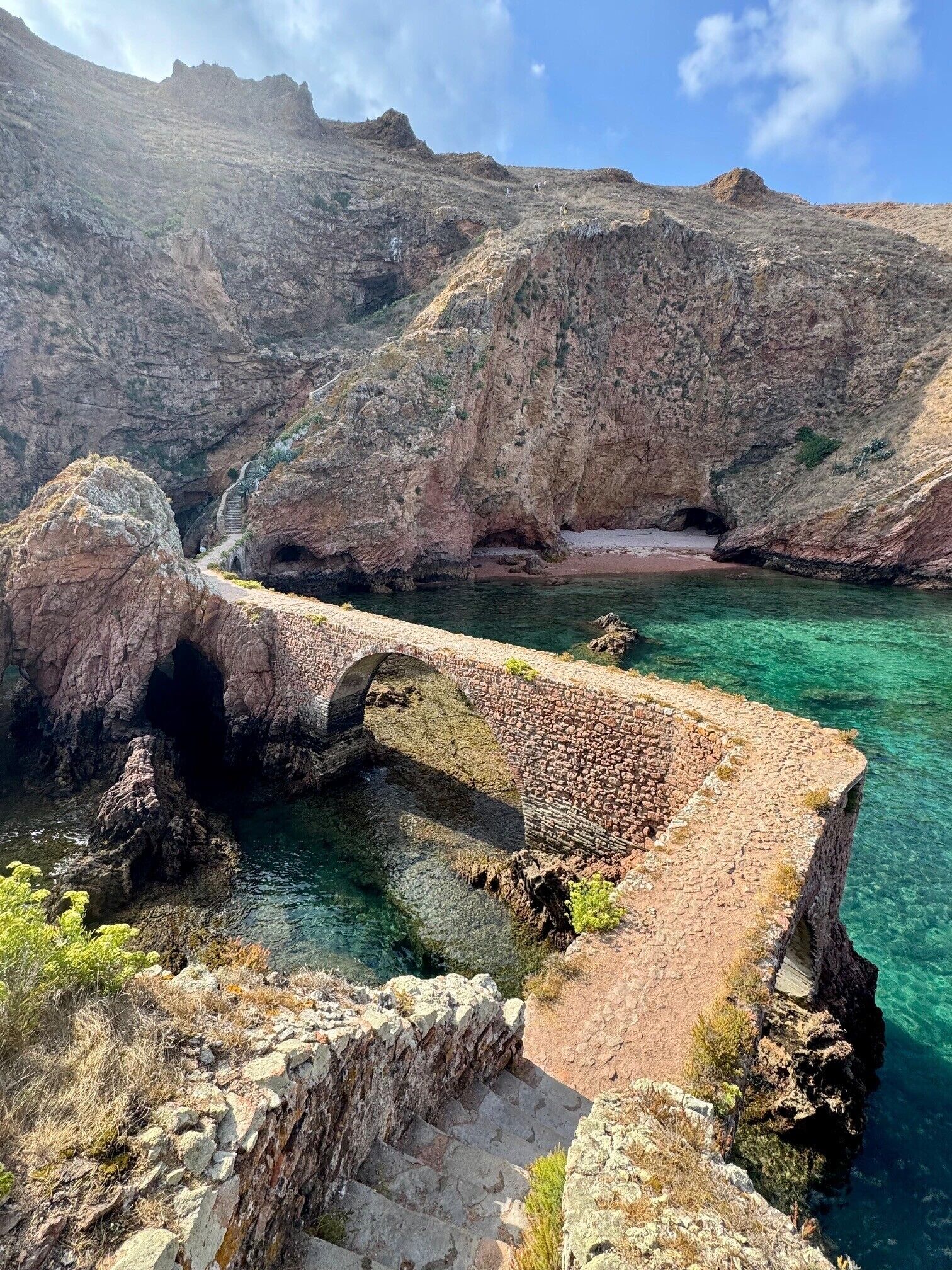 A stone bridge over a clear blue water body, surrounded by rocky terrain.
