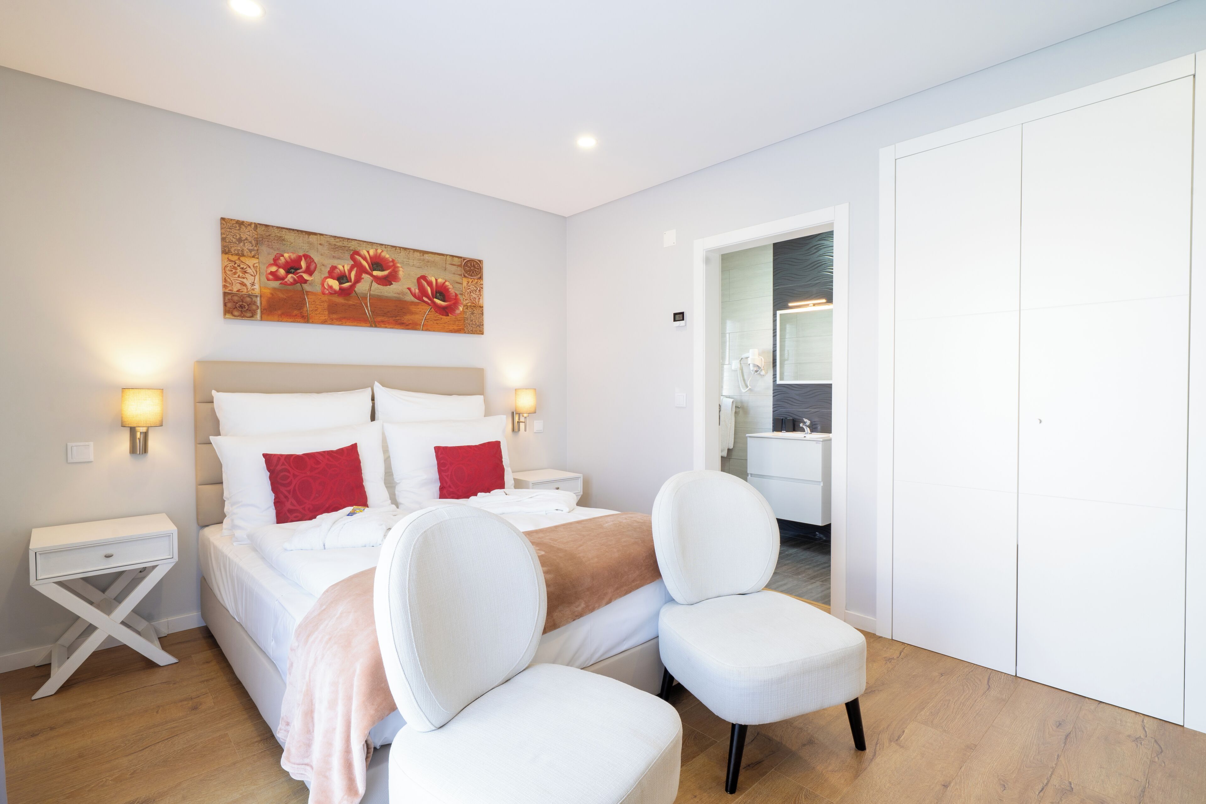 A hotel room with a bed, bedside tables, a chair, a painting, and a bathroom visible through an open door.