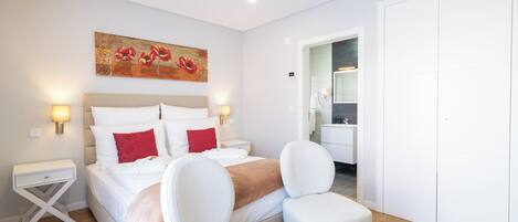 A hotel room with a bed, bedside tables, a chair, a painting, and a bathroom visible through an open door.