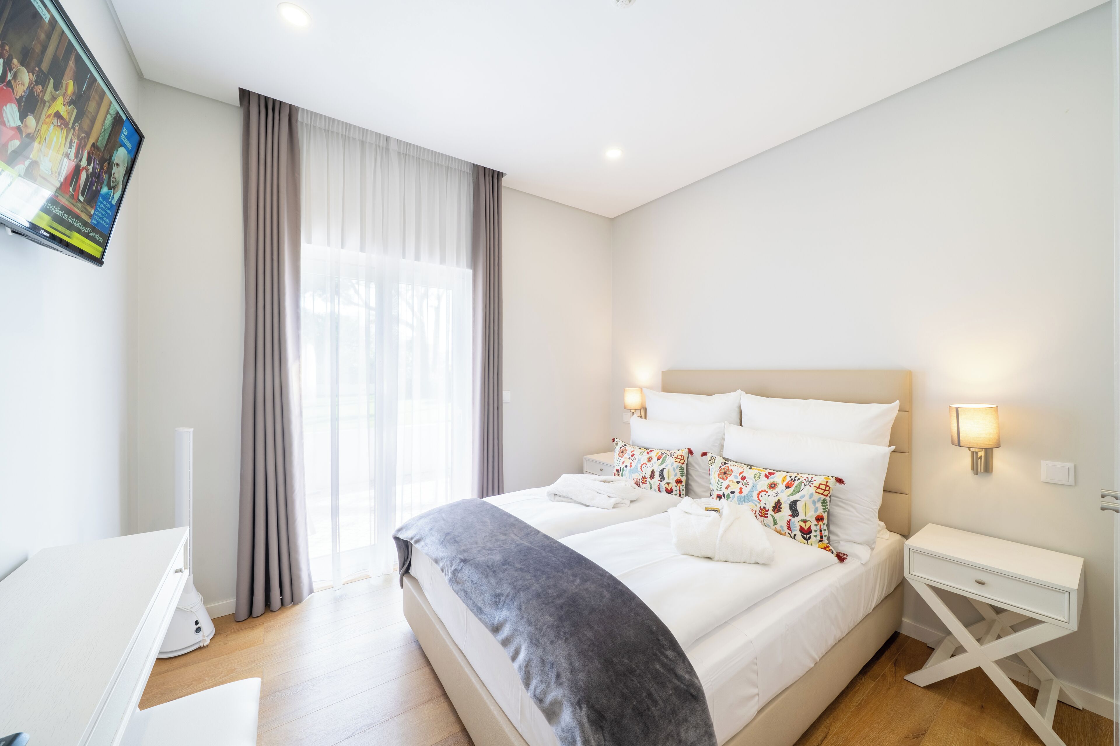 A modern hotel room with a large bed, bedside tables, a flat-screen TV, and a large window with curtains.