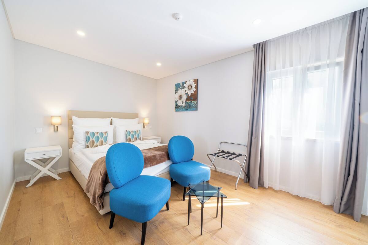 Een hotelkamer met een bed, twee blauwe stoelen, een kleine tafel, een wandlamp en een raam met gordijnen.