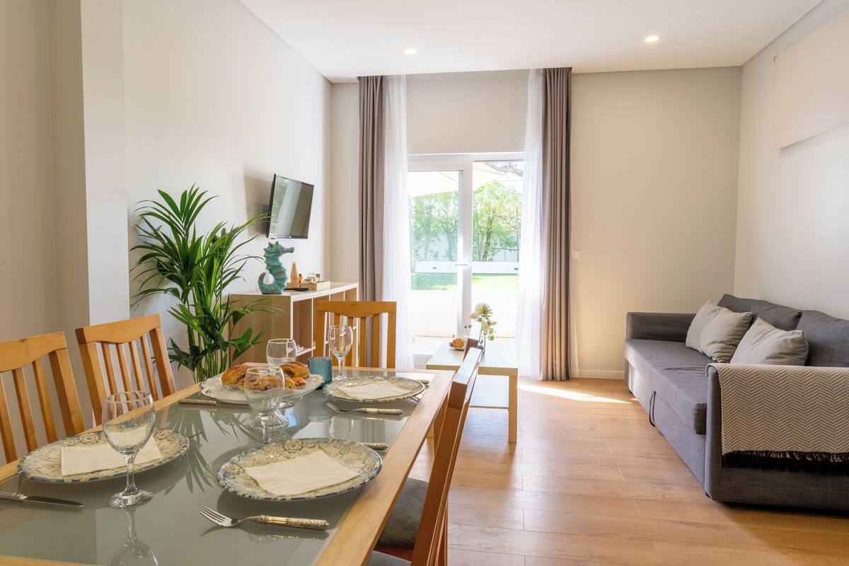Een eethoek met een glazen tafel voor vier personen, een bank, een televisie aan de muur en een plant.