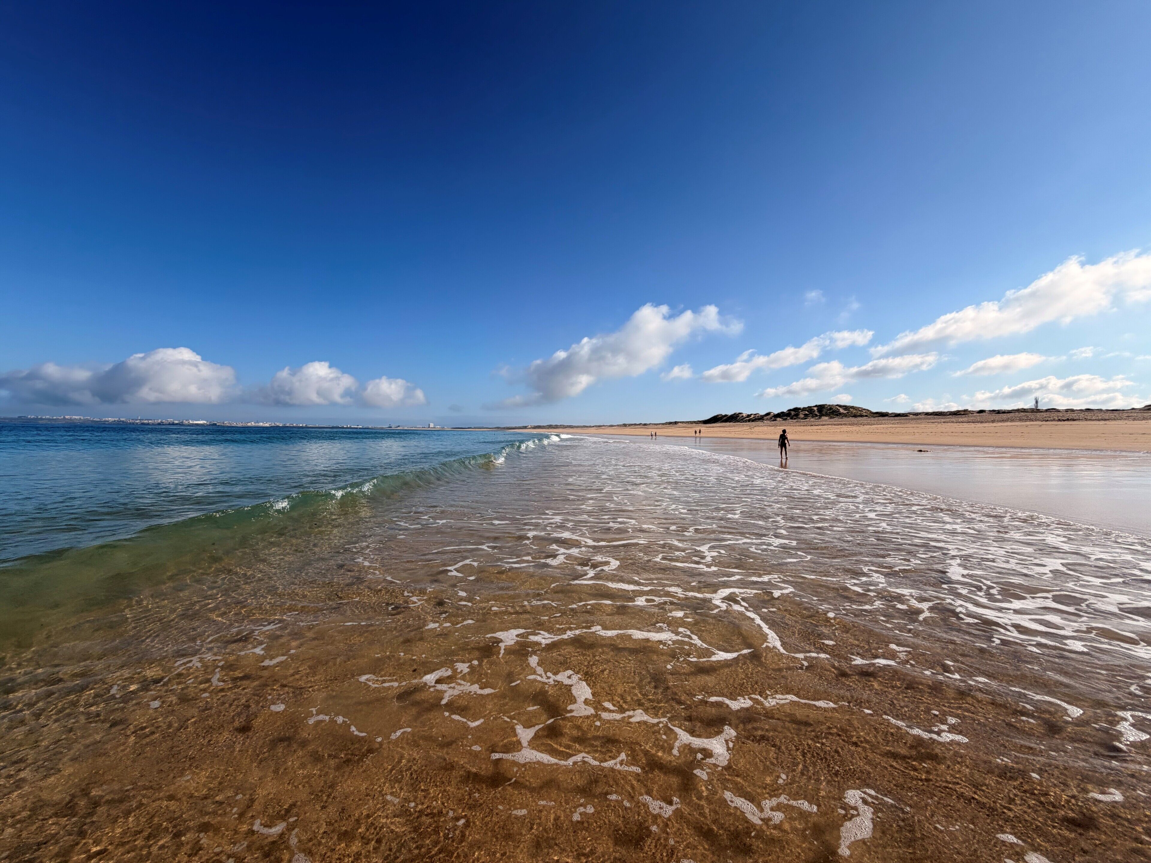 A beach with clear water, a person walking, and distant buildings.