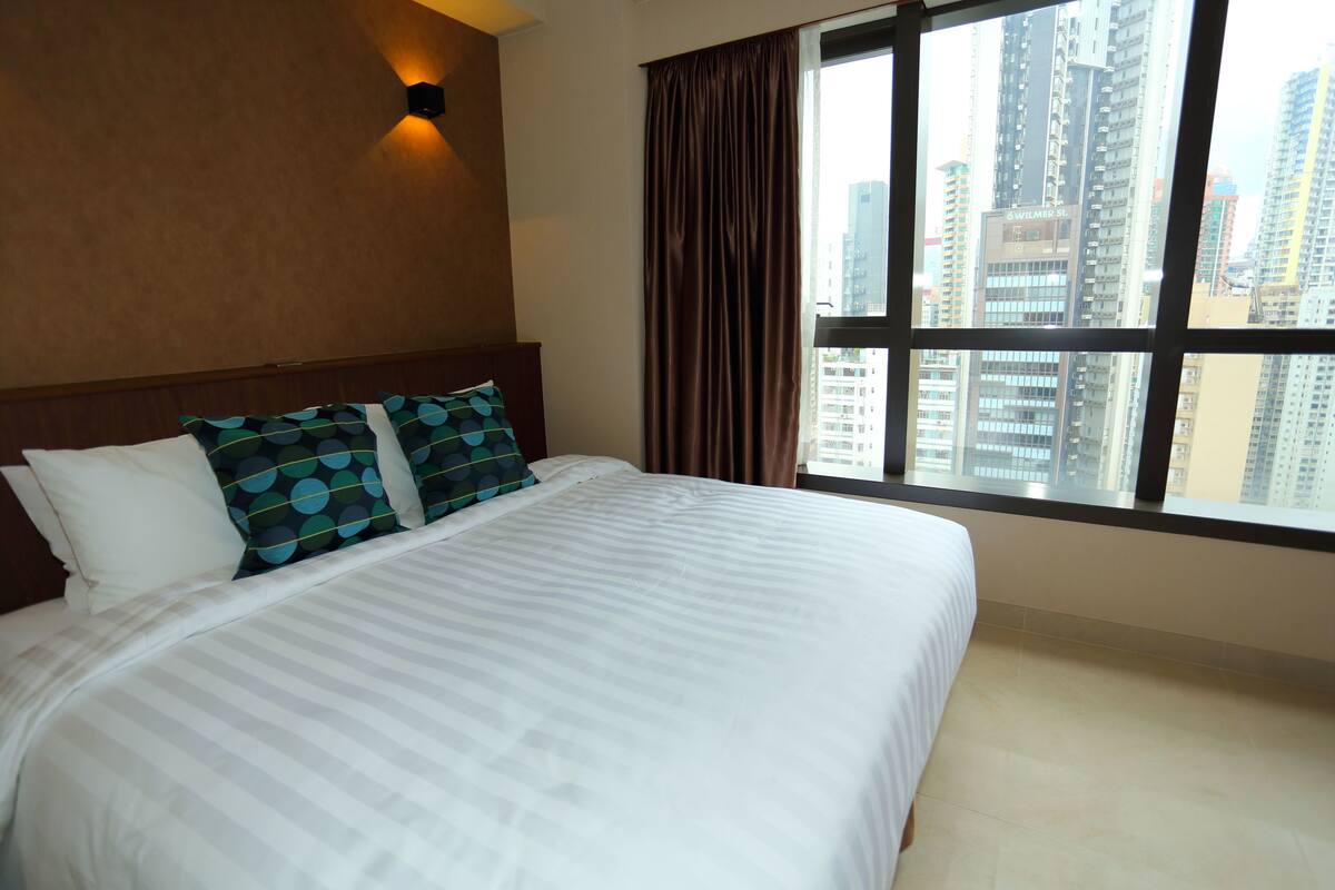 A hotel room with a large bed, a view of tall buildings, and a window with curtains.