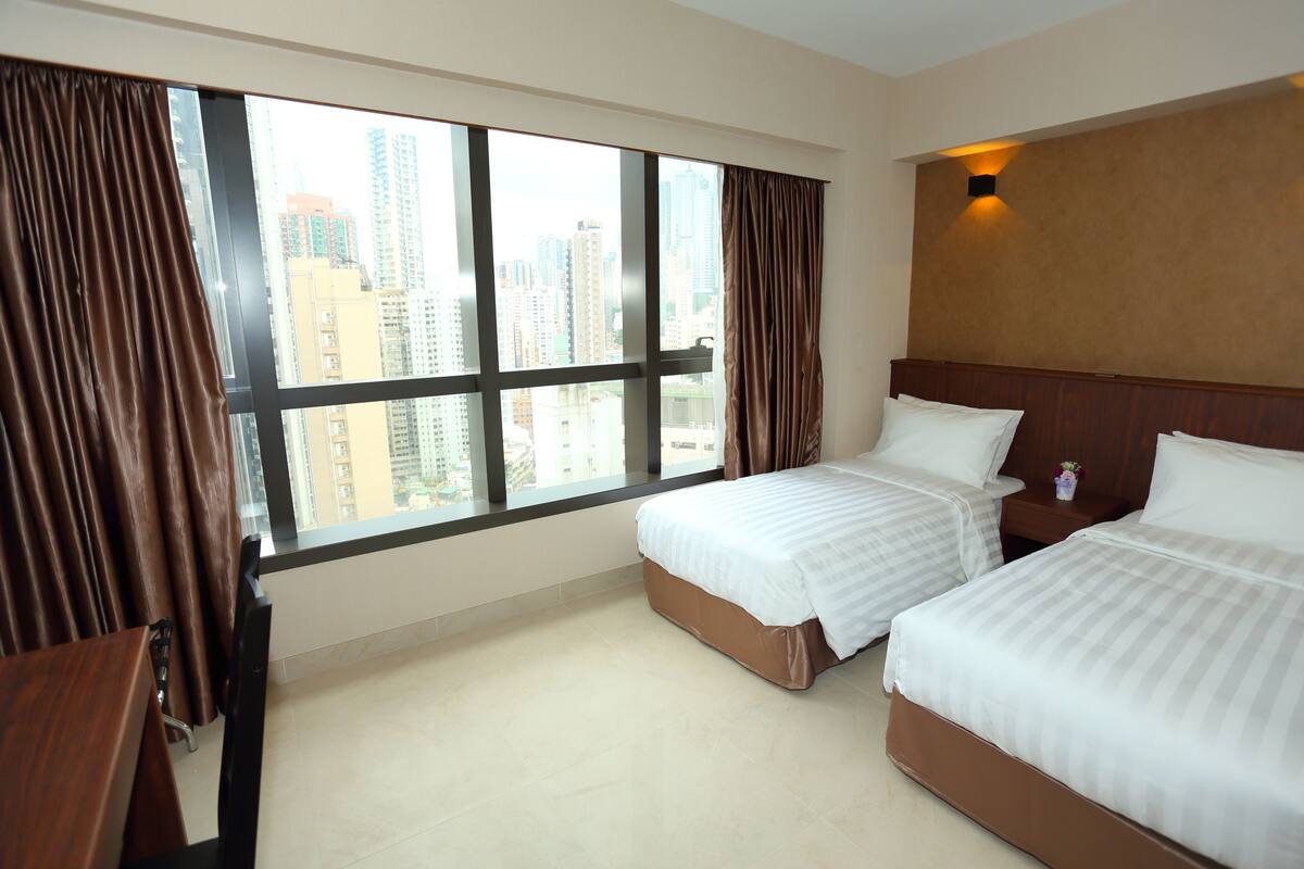 A hotel room with two beds, a large window with curtains, a wooden headboard, and a nightstand with a vase.