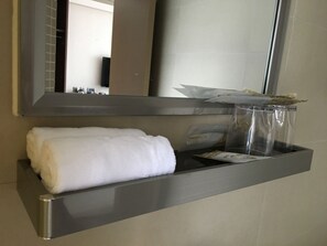 A bathroom with a towel rack holding a rolled towel, a glass container with toiletries, and a mirror reflecting the room. - WE Hotel (Hong Kong)