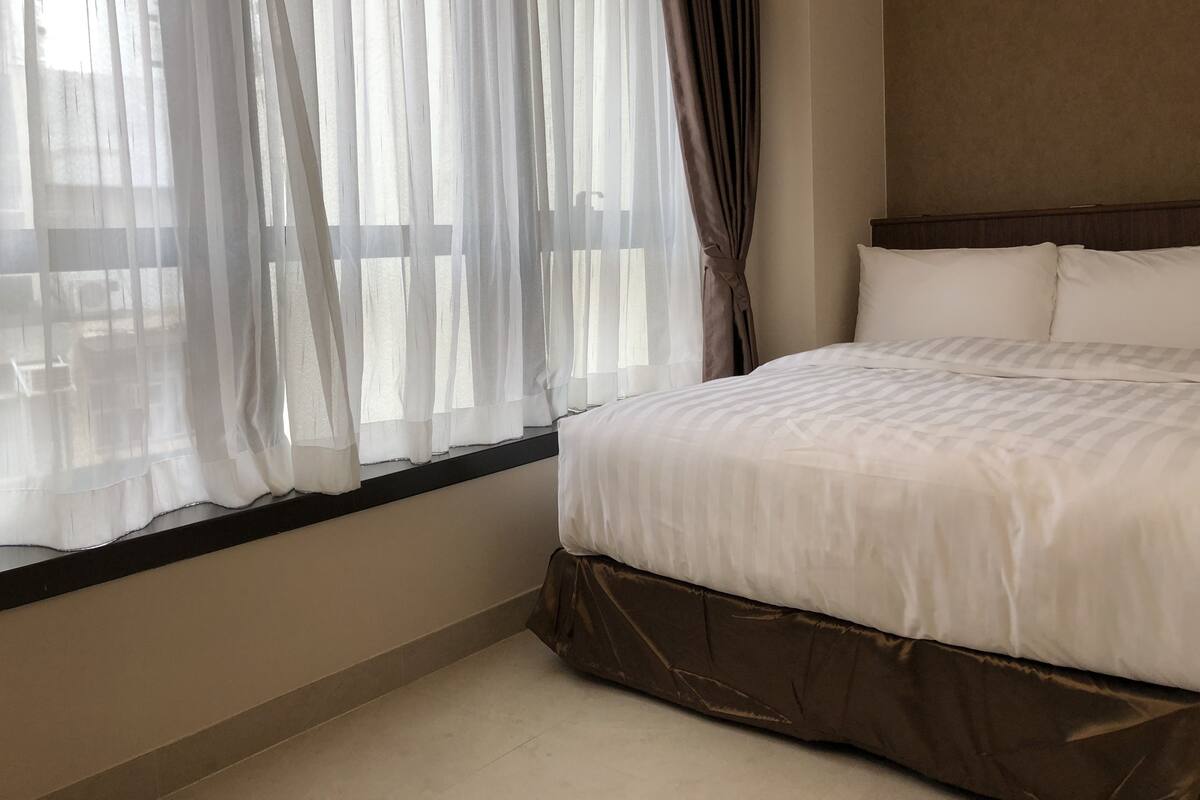 A hotel room with a large bed, a window with sheer curtains, and a brown headboard.