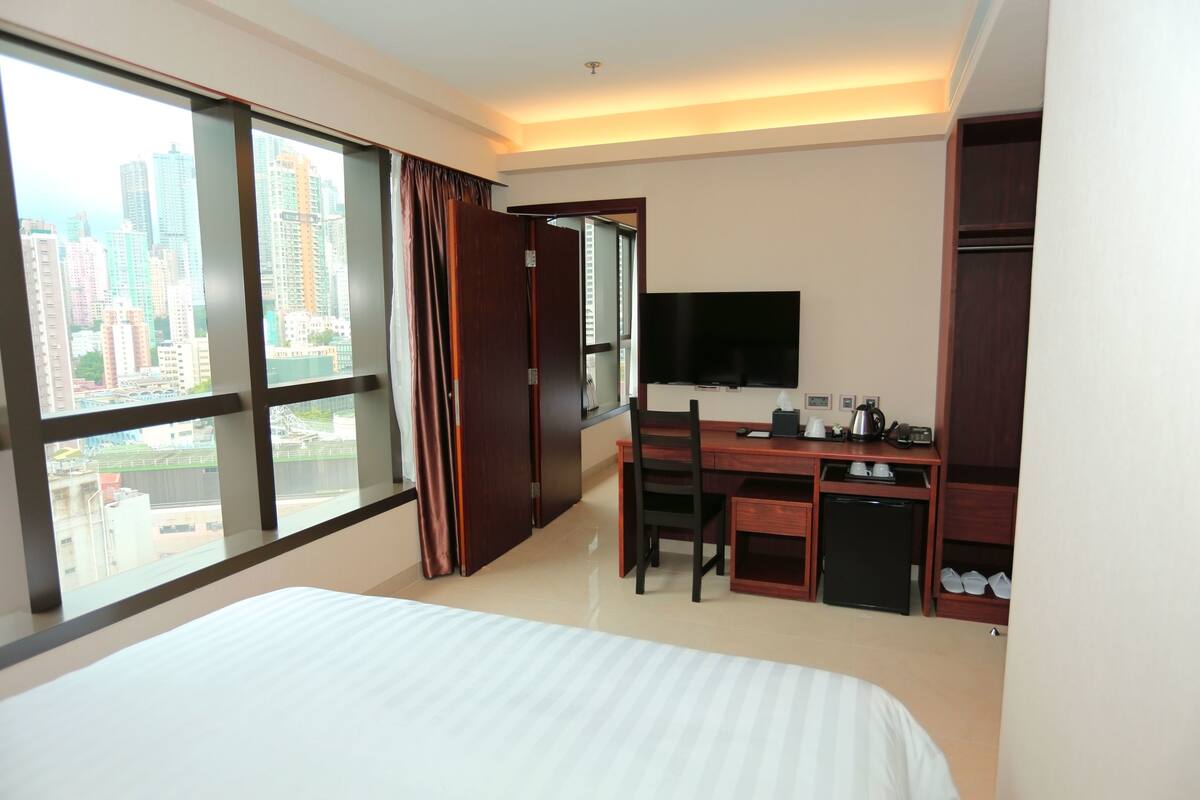 A hotel room with a large bed, a desk with a chair, a television, and a view of the city through large windows.