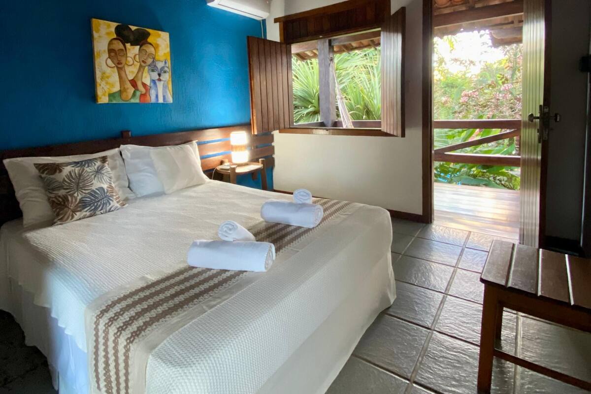 A bedroom with a bed, pillows, a painting, an air conditioning unit, a window, and a door leading outside.