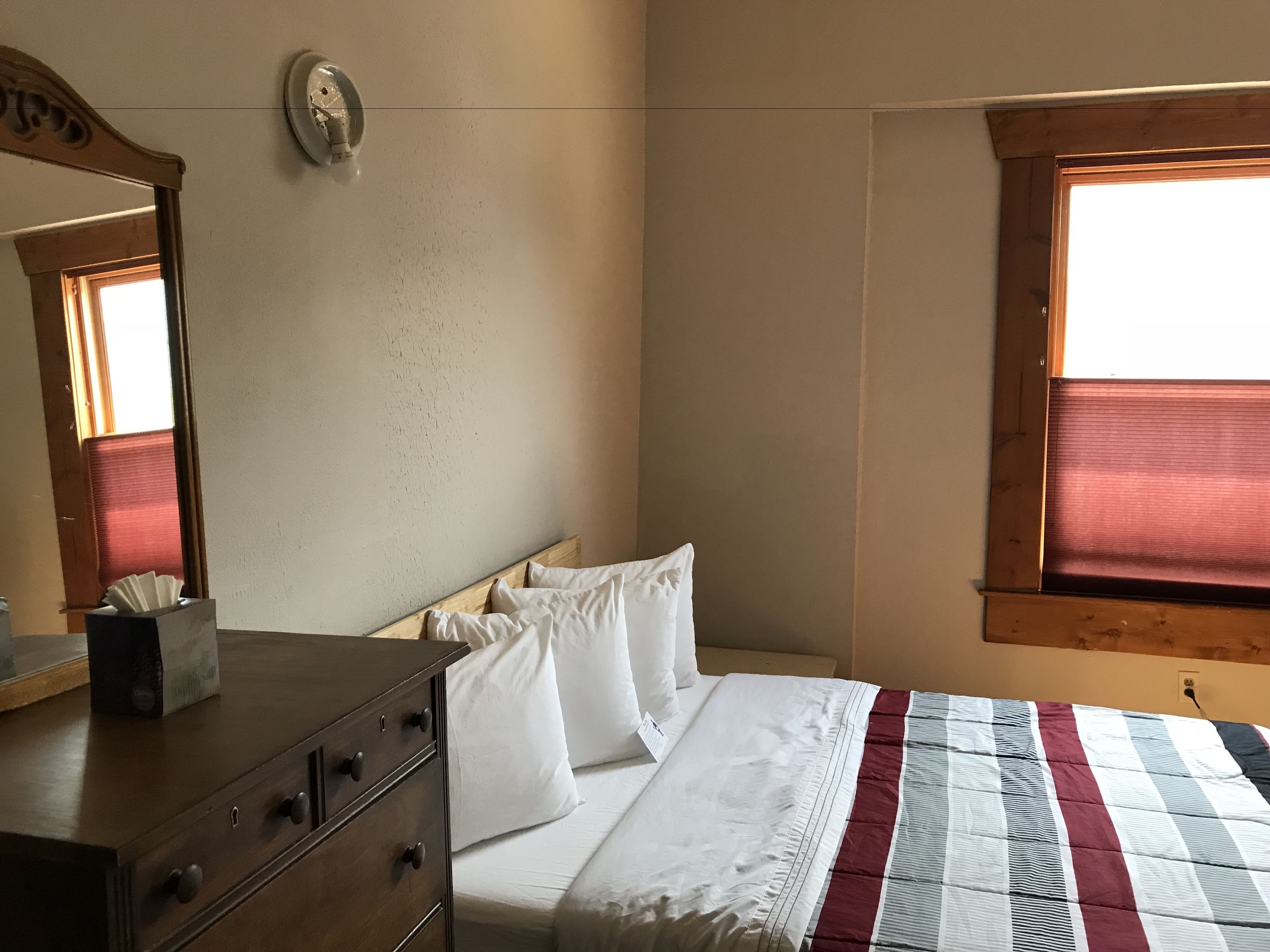 Un dormitorio con un aparador de madera, una cama con ropa de cama blanca, un ventanal con persianas y un espejo montado en la pared.