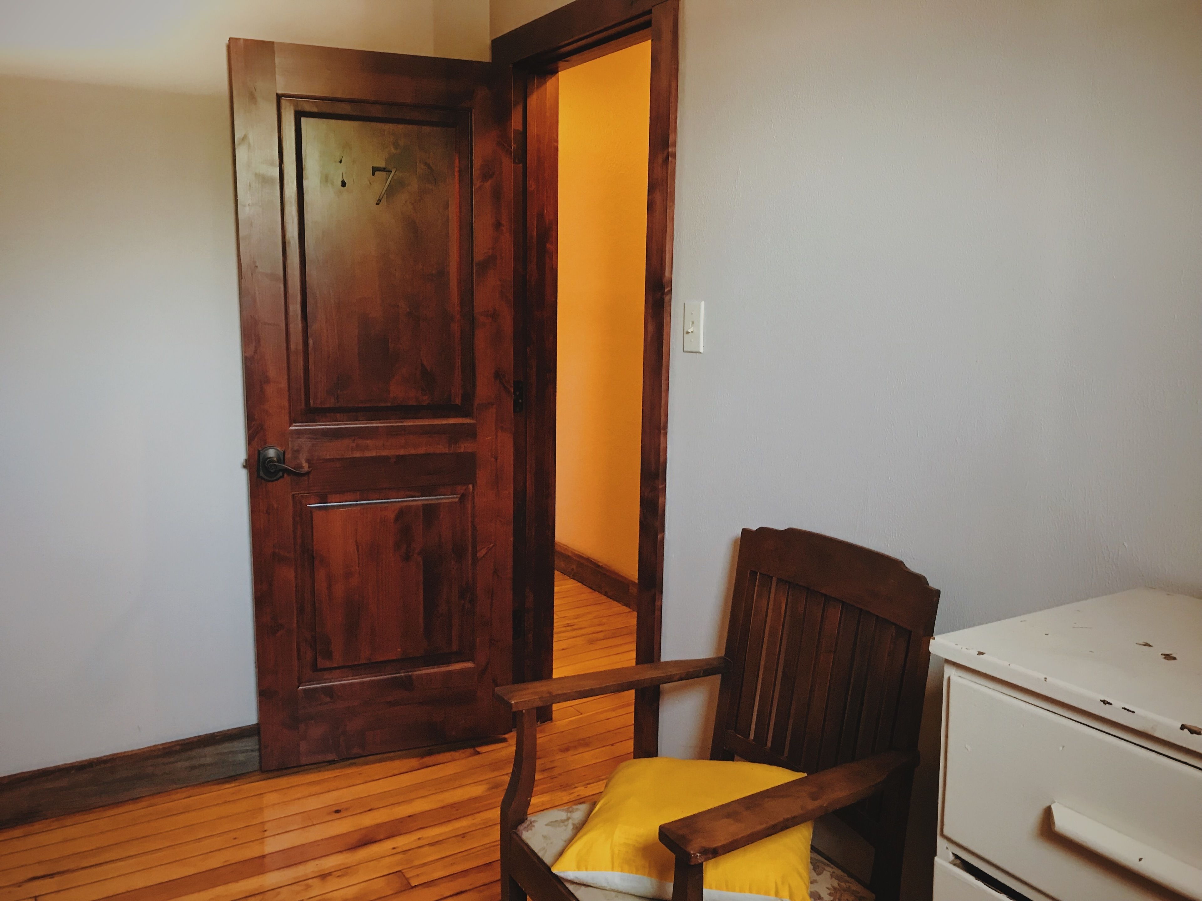 A room with a wooden door, a wooden chair with a yellow cushion, and a white dresser.