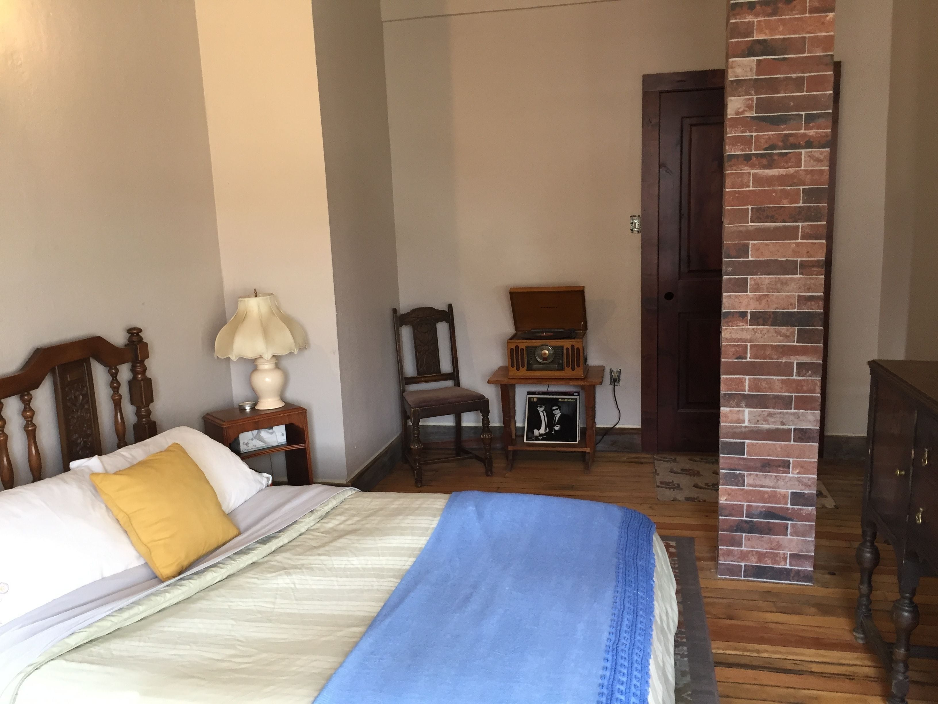 Un dormitorio con una cama, una mesita de noche, una lámpara, una silla, una pequeña mesa con una radio y un pilar de ladrillo.