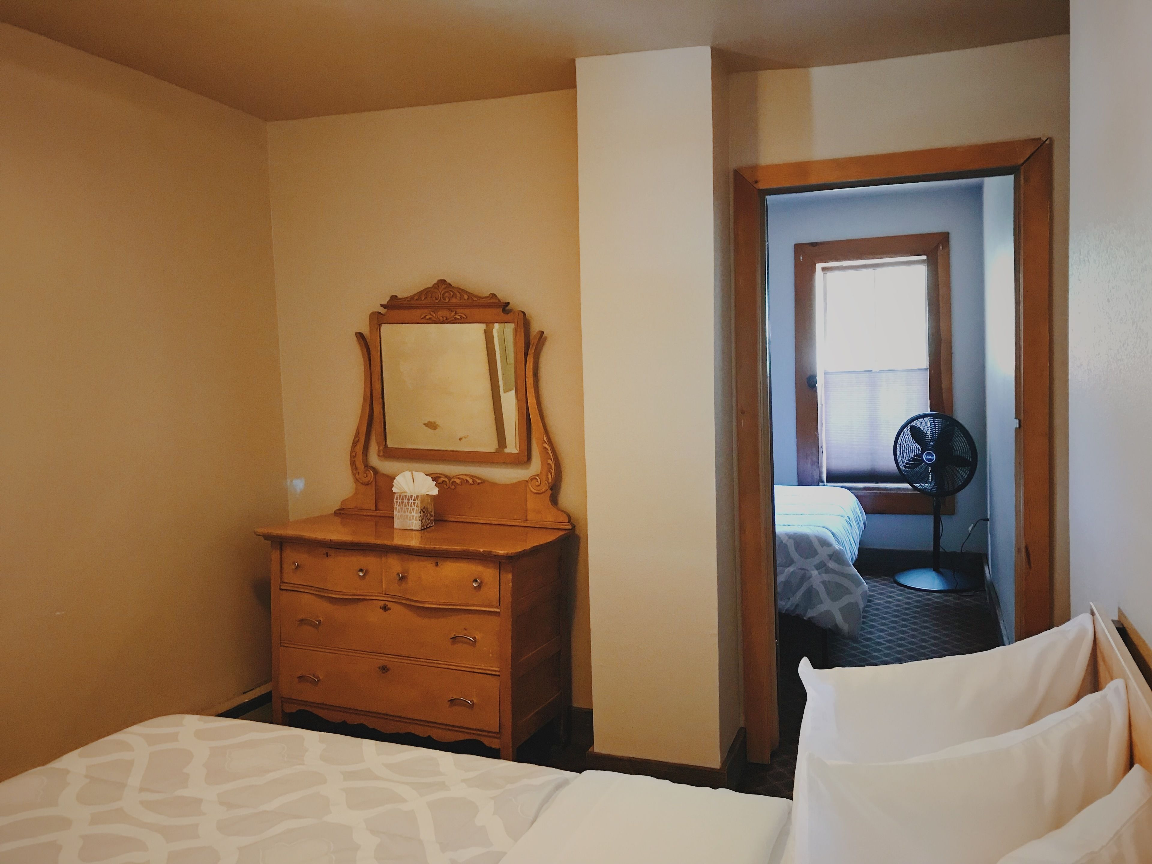 A hotel room with a bed, a wooden dresser with a mirror, and a door leading to another room.