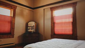 A bedroom with a large bed, wooden headboard, a dresser, a mirror, and two windows with blinds.