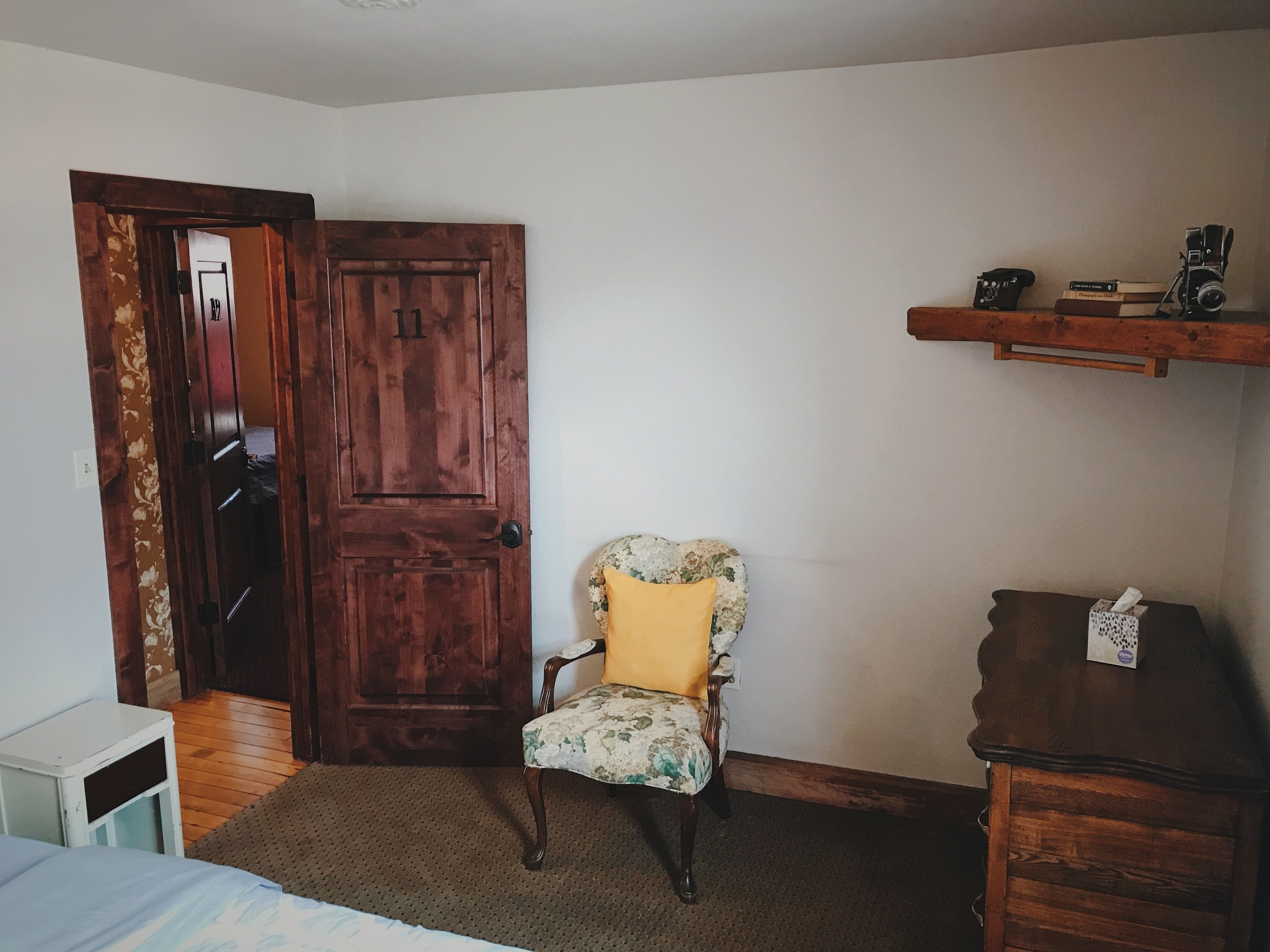 Una habitación con puerta de madera, un sillón tapizado con flores y un cojín amarillo, una pequeña mesa blanca y un aparador de madera con un dispensador de pañuelos encima.