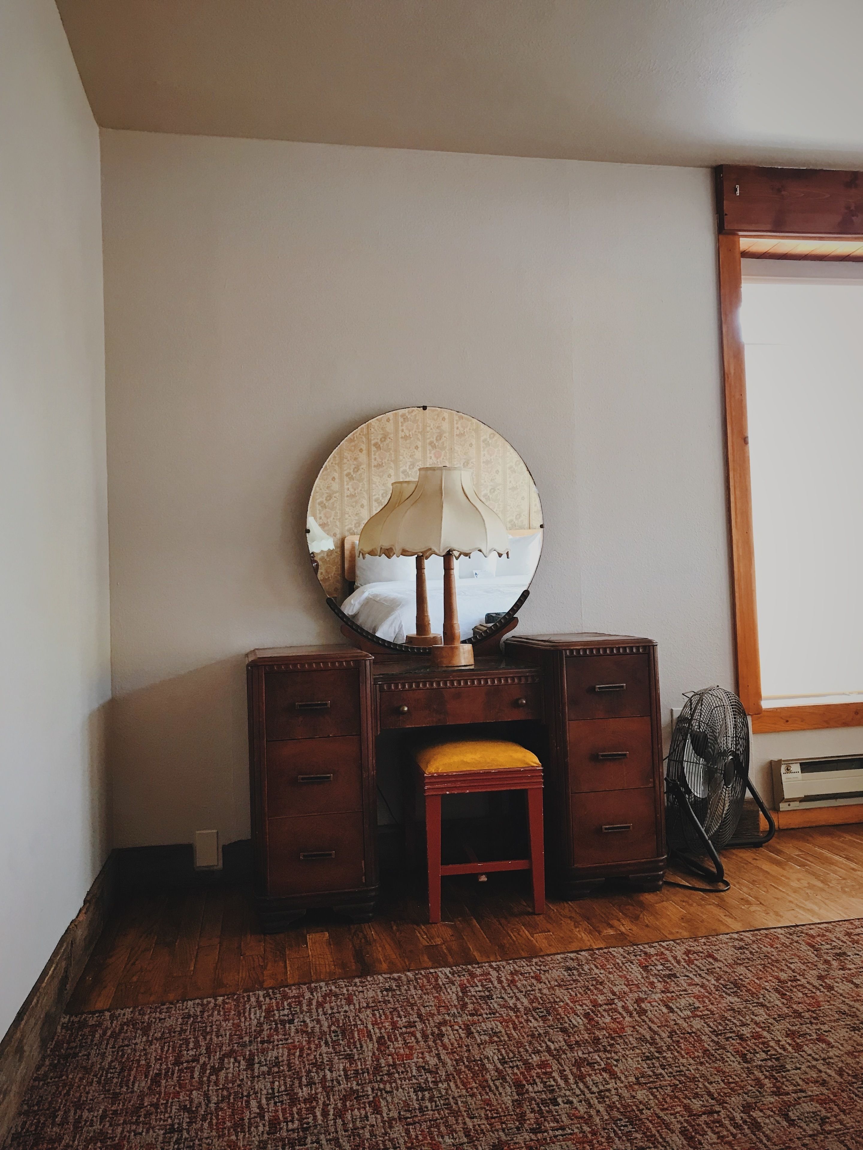 Un aparador de madera con espejo y un taburete en una habitación con piso de madera y un tapete estampado.