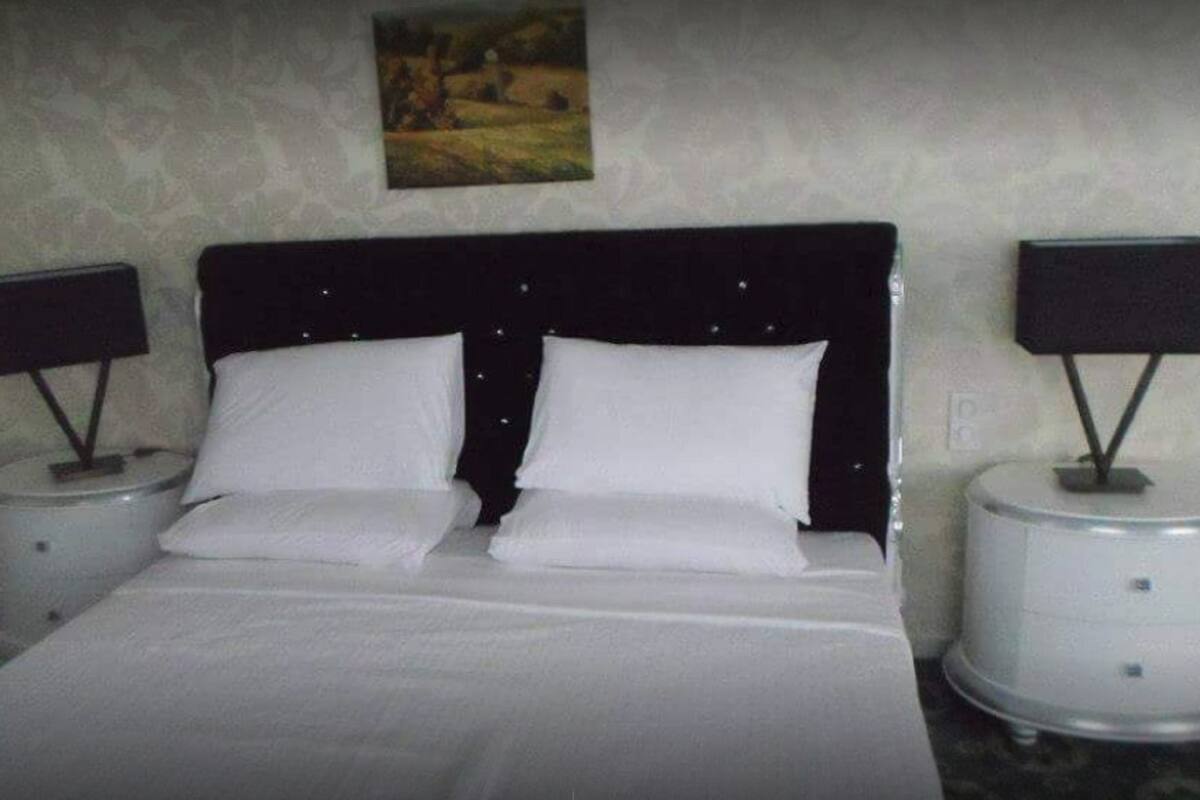 Una cama bien hecha con sábanas blancas y dos almohadas, flanqueada por dos mesitas de noche con lámparas, en una habitación con pared texturizada y un cuadro de paisaje sobre la cama.