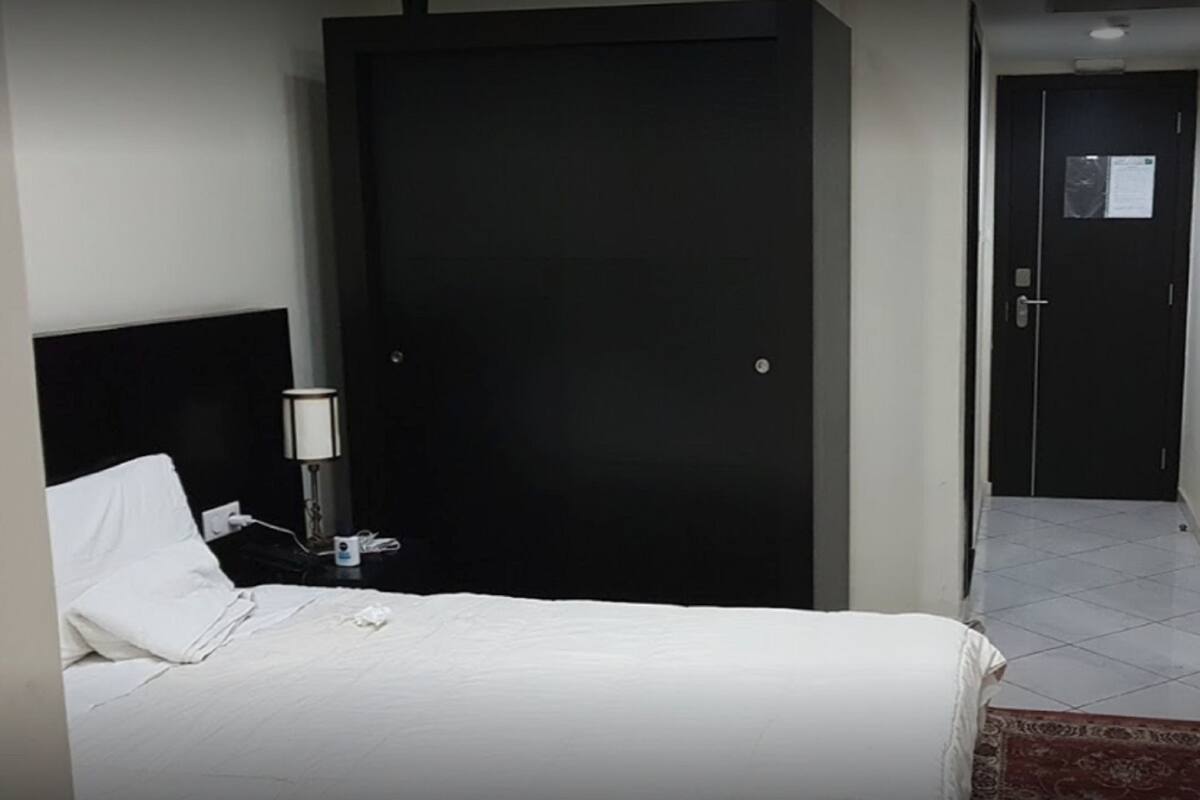 Habitación de hotel con cama, mesita de noche, lámpara, televisor y puerta que da a un pasillo.
