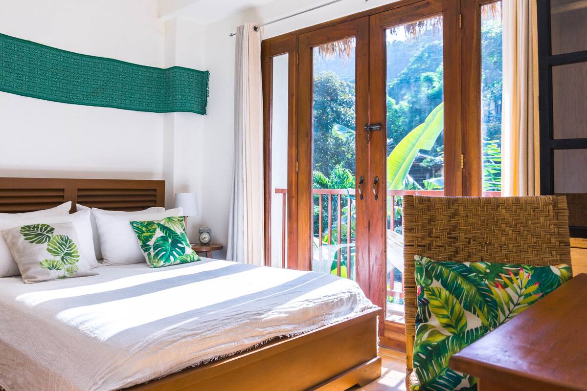 Une chambre à coucher avec un lit, des oreillers, une table de chevet, une lampe et une vue sur un jardin tropical, visible à travers la porte.