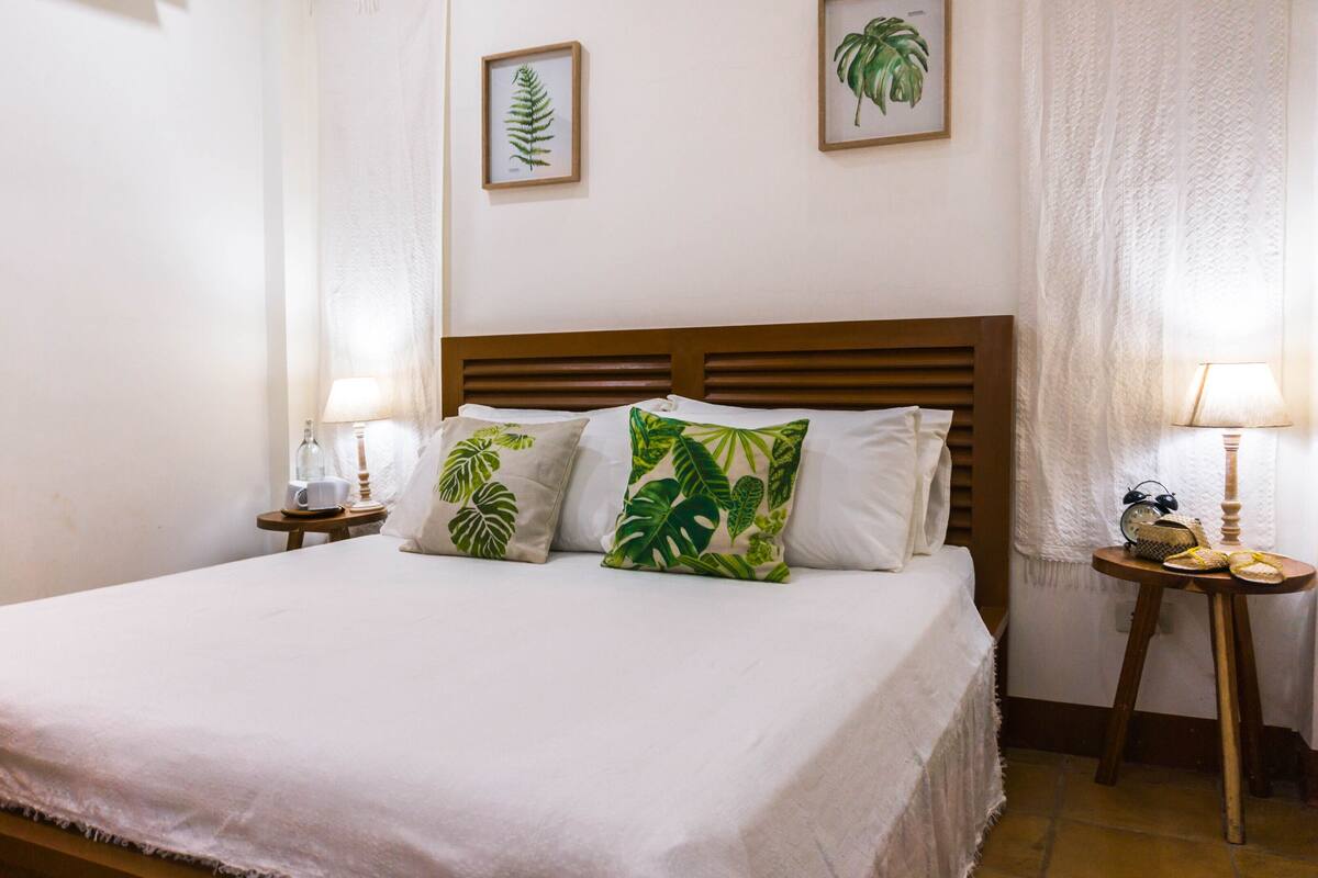 Un lit bien fait, avec du linge de lit blanc, deux tables de chevet avec des lampes et des coussins ornés de motifs tropicaux.
