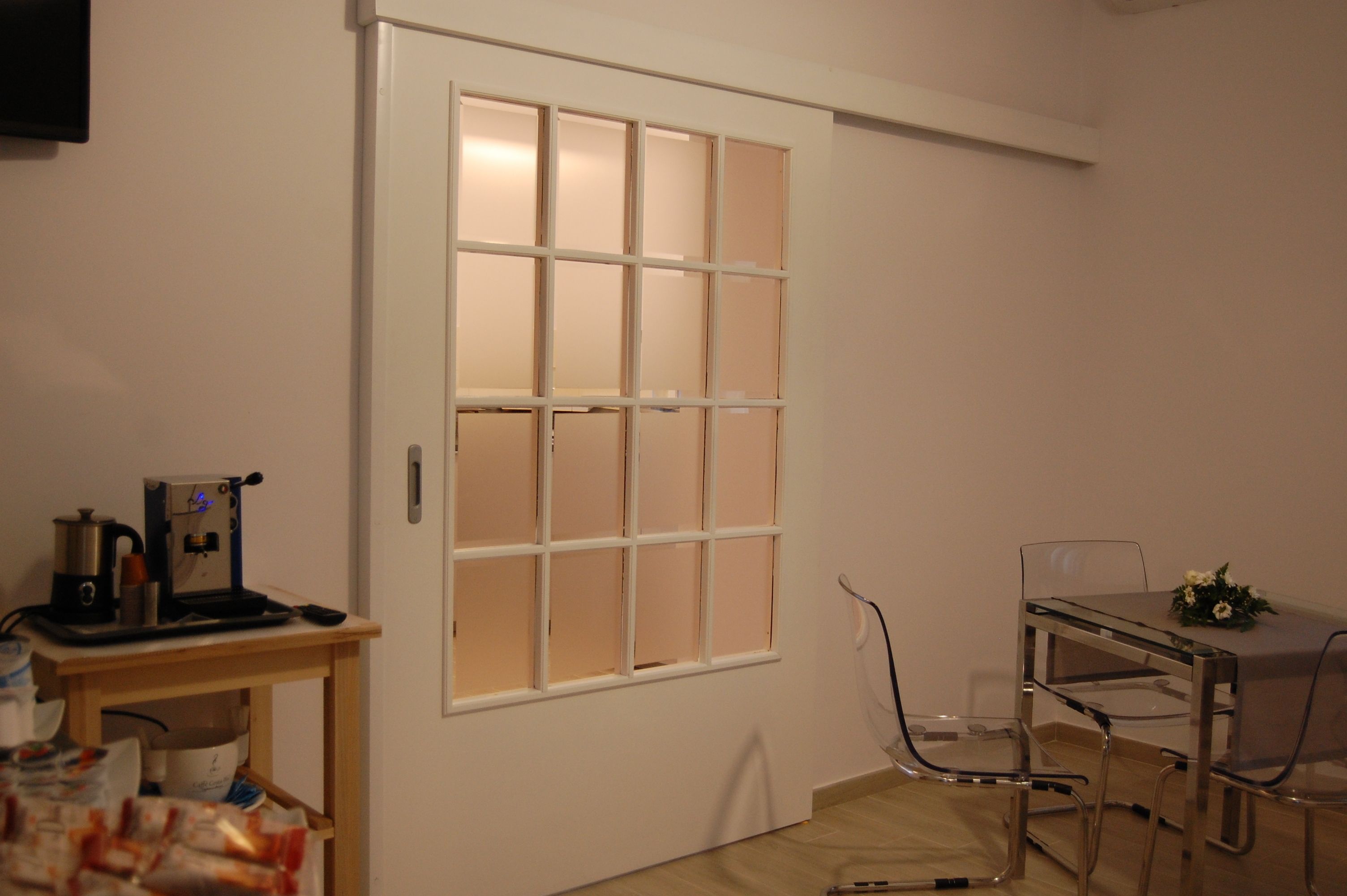 A room with a white door, a small table with a coffee machine, and a glass table with chairs.