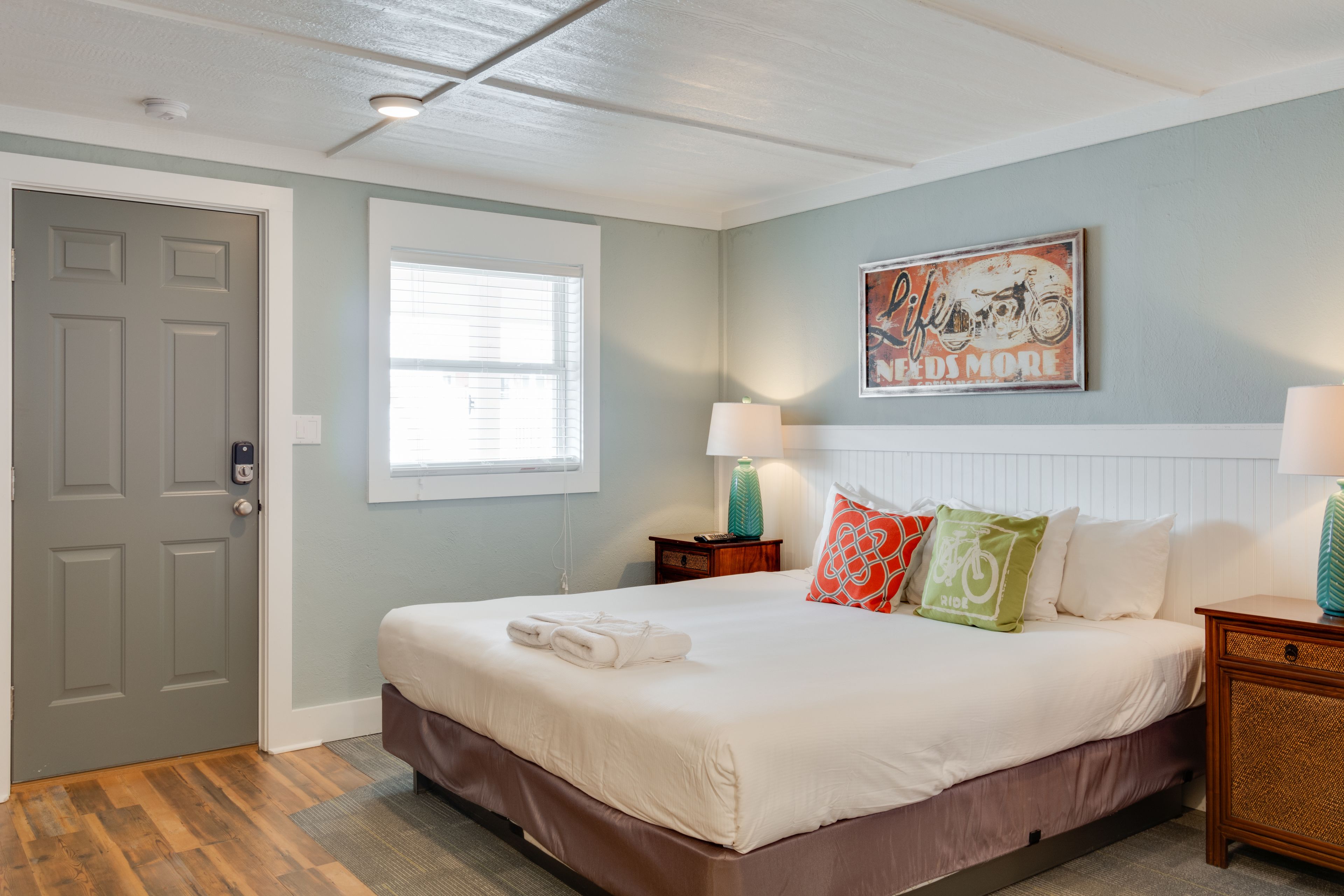 A bedroom with a bed, a door, a window, a wall-mounted picture, and a nightstand with a lamp.