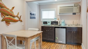 Cocina compacta con una mesa de madera, sillas blancas, armarios oscuros, un microondas, una cafetera y un aire acondicionado.