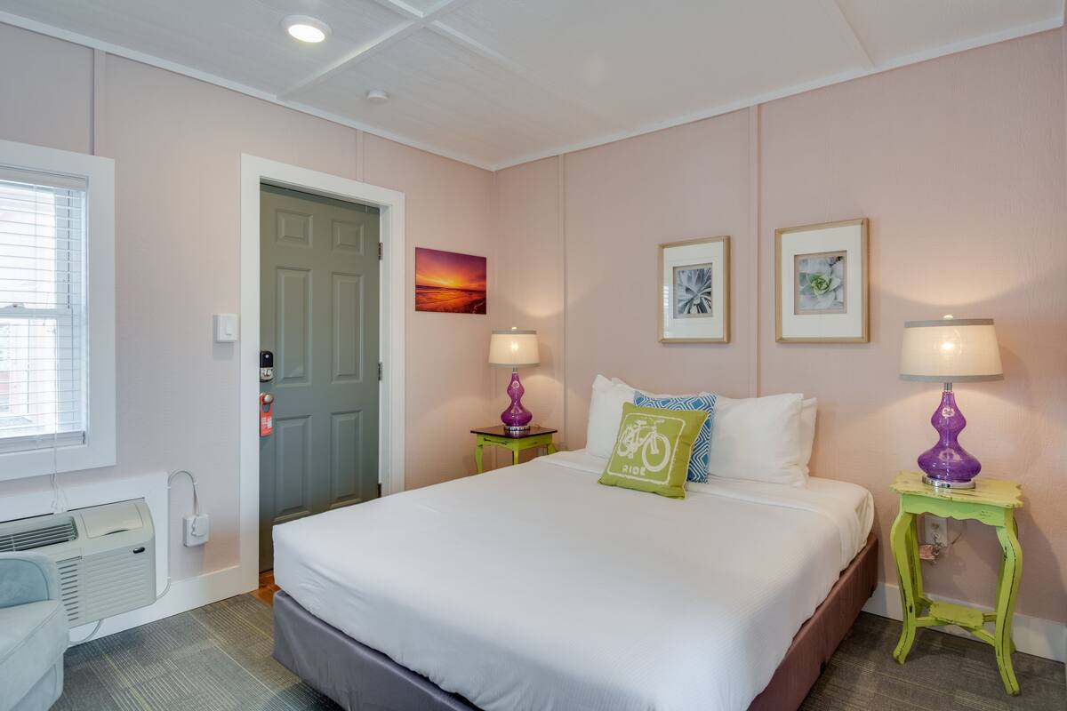 A neatly arranged hotel room with a bed, two bedside tables with lamps, a window with blinds, and a door.