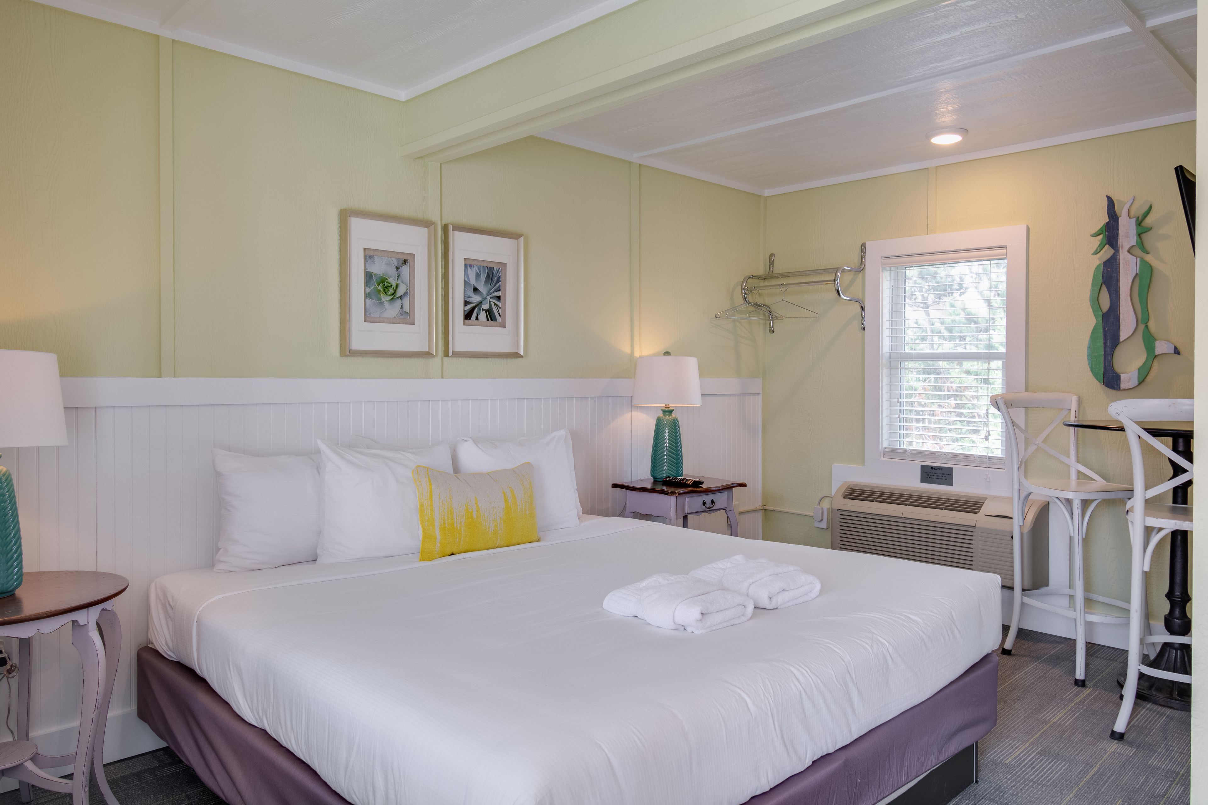 A hotel room with a large bed, a bedside table with a lamp, a chair, a window with blinds, and artwork on the wall.