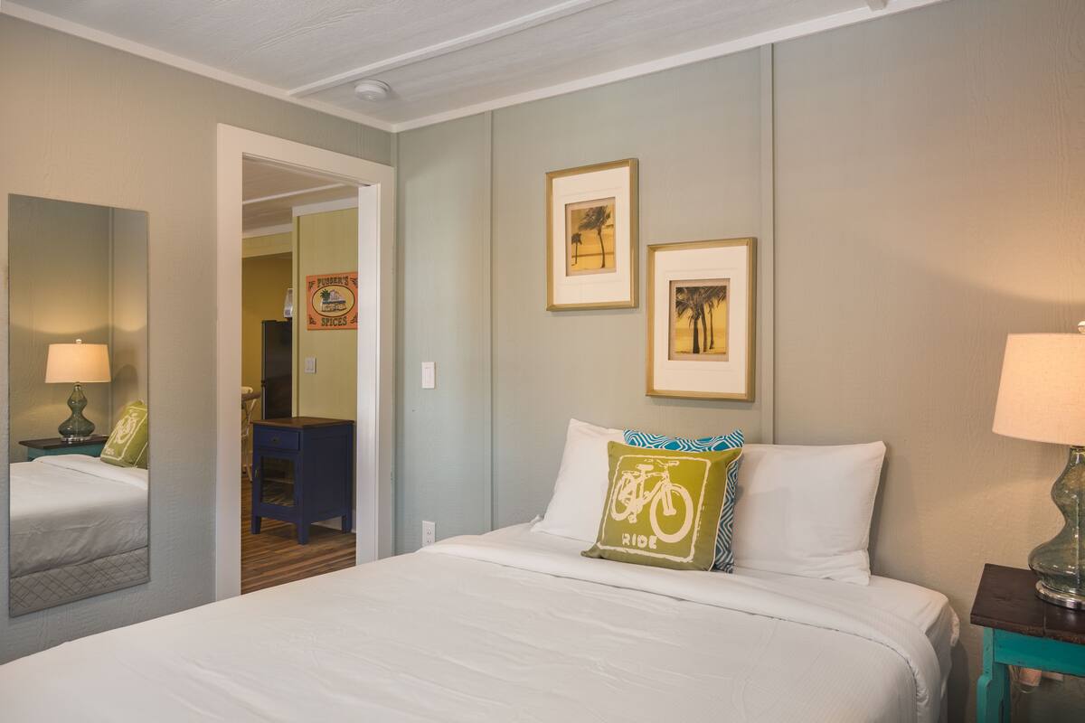 A hotel room with a bed, a nightstand with a lamp, a mirror, and two framed pictures on the wall.