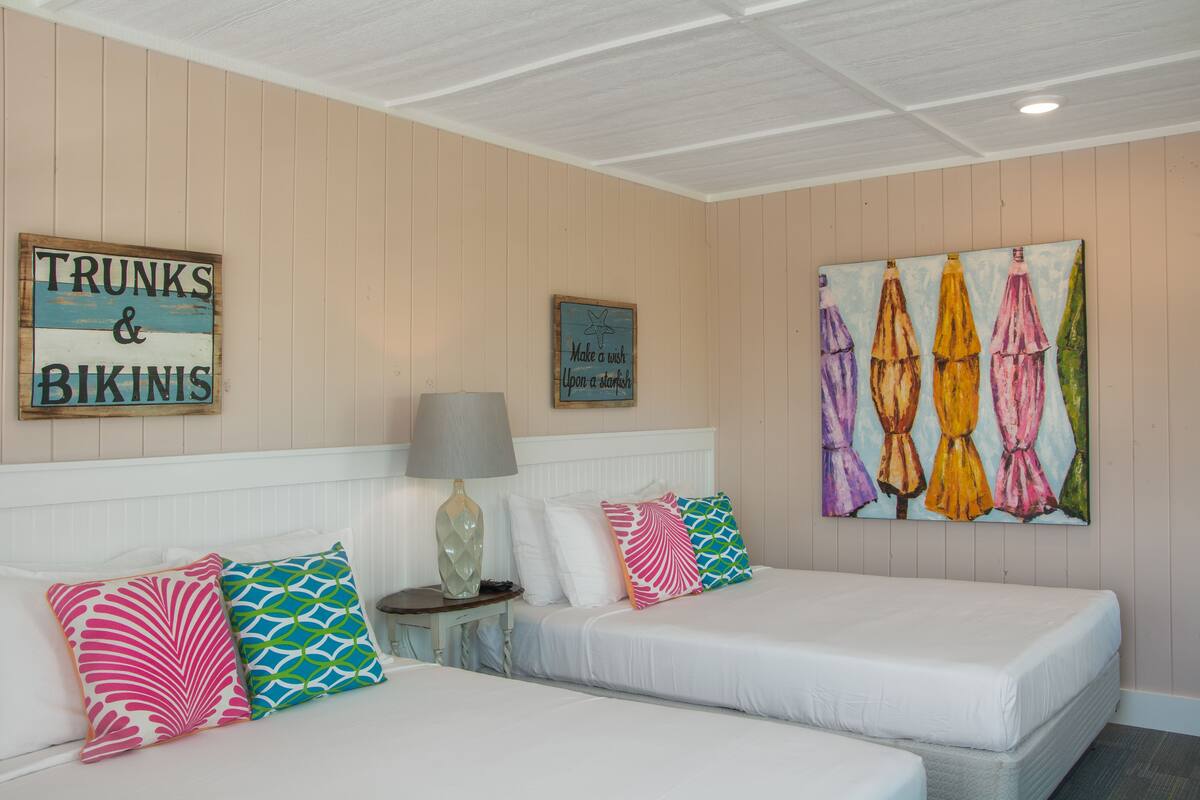 A hotel room with two beds, a nightstand, a lamp, and wall art including a sign that reads "TRUNKS & BIKINIS" and a painting of colorful umbrellas.