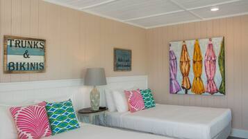 A hotel room with two beds, a nightstand, a lamp, and wall art including a sign that reads "TRUNKS & BIKINIS" and a painting of colorful umbrellas.