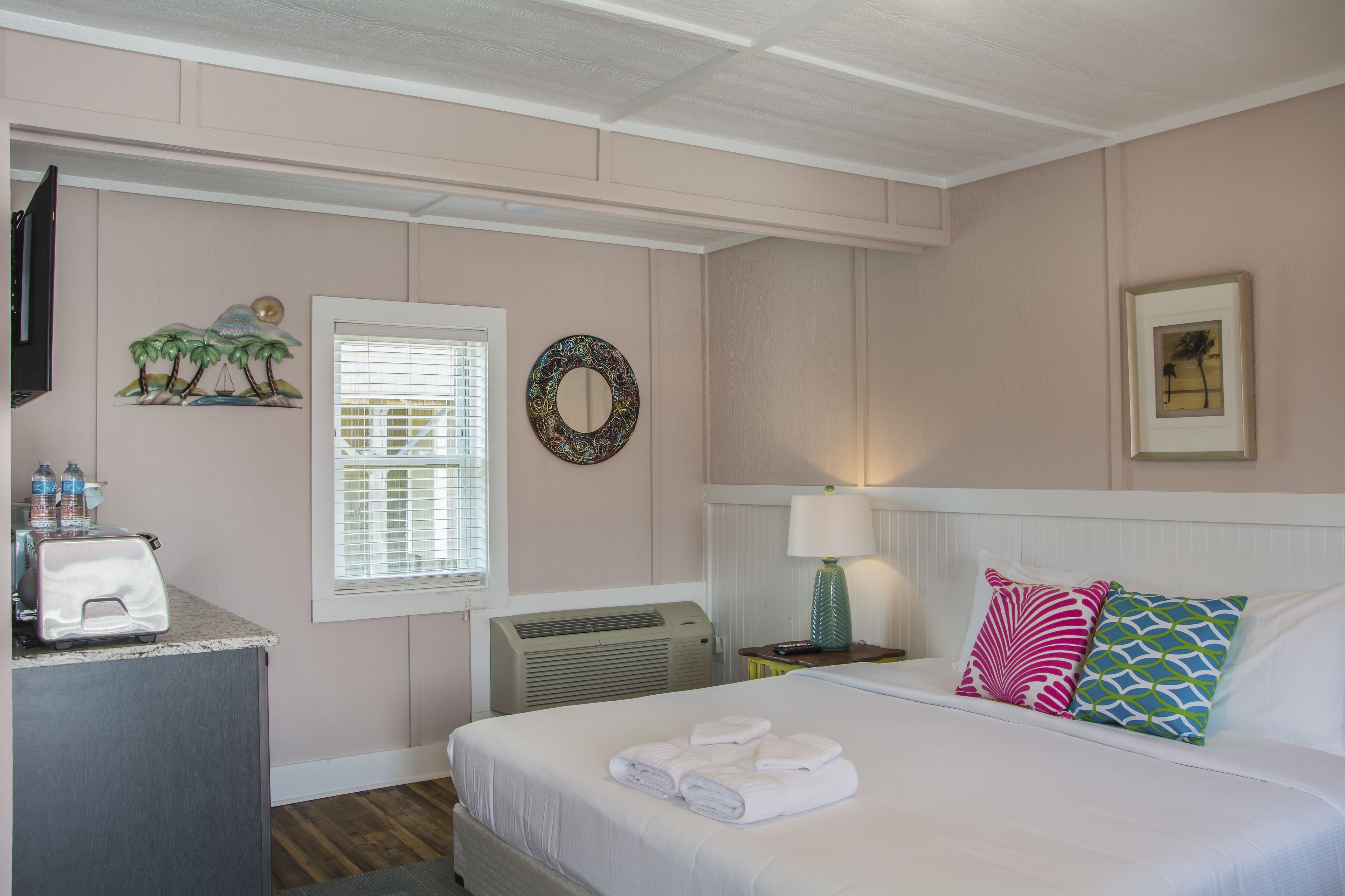 Quarto de hotel com cama, escrivaninha com torradeira, janela com persianas, espelho fixado na parede e quadro emoldurado.