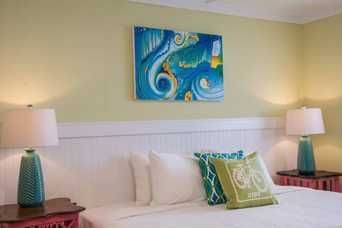 A hotel room with a bed, white headboard, two nightstands with lamps, and a colorful abstract painting on the wall.