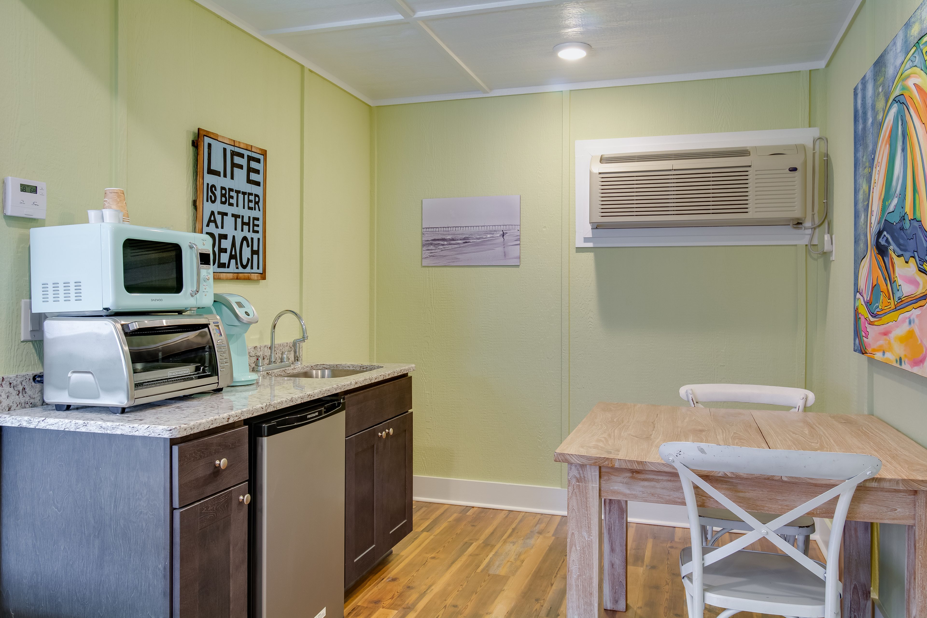 A compact kitchen with a microwave, toaster, and sink. There's a wooden table and a chair, and an air conditioner mounted on the wall.