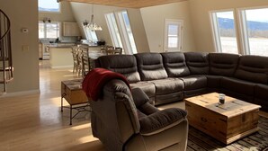 Un salon spacieux avec un grand canapé d’angle, une table basse en bois et une vue sur la cuisine et la salle à manger grâce à de grandes fenêtres.