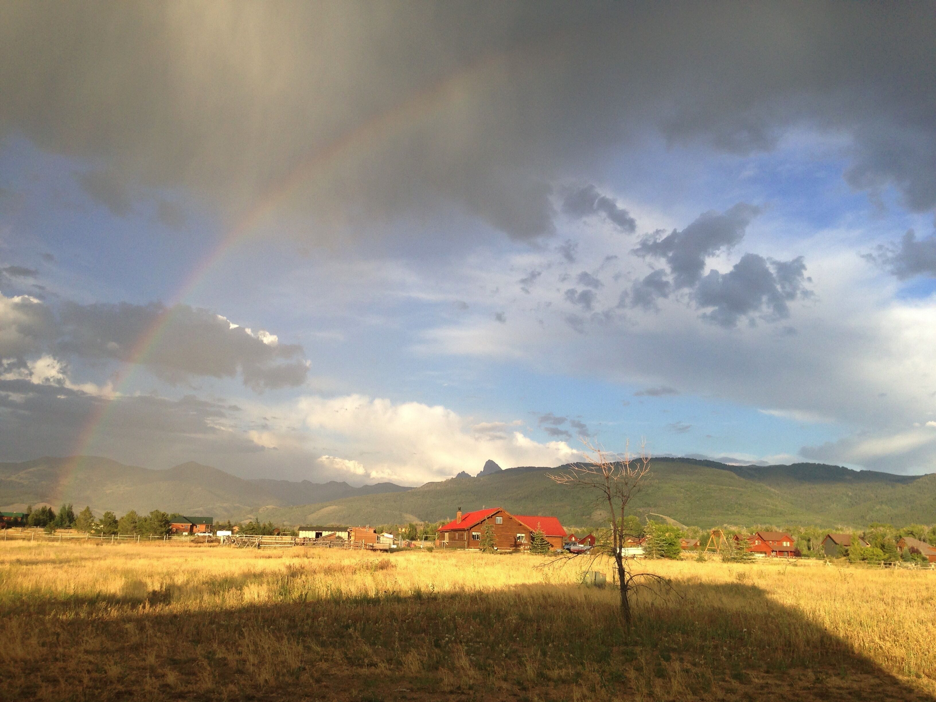 A rural landscape with a rainbow, houses, and mountains in the background.