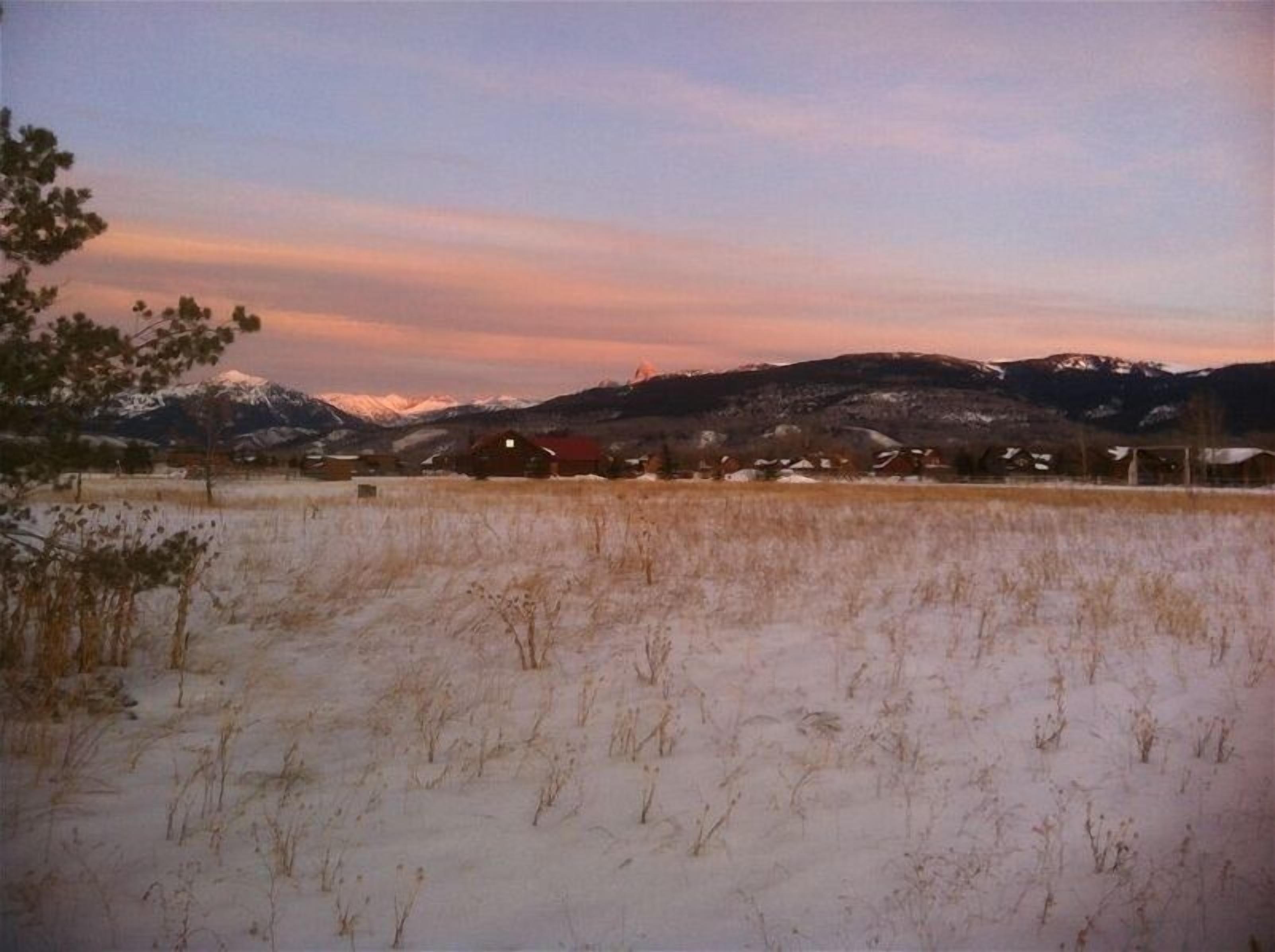 Ett lantligt landskap med snötäckta fält, några hus och berg i bakgrunden.