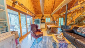 Un interior acogedor de cabaña con comedor, sala de estar con sofá, chimenea y ventanas amplias que ofrecen vistas al exterior.