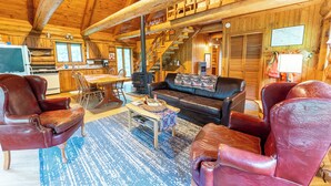 Interior de una cabaña de troncos con comedor, sala de estar con muebles de cuero y una escalera que conduce a un nivel superior.