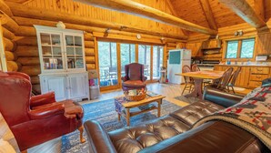 Interior de una cabaña de troncos con una sala de estar que incluye un sofá de cuero, un sillón rojo y una mesa de centro de madera. Se ve un mueble blanco con puertas de vidrio, y un área de cocina con estufa y refrigerador al fondo.