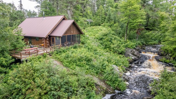 Una cabaña de troncos con techo metálico, rodeada de un bosque frondoso y una cascada pequeña.