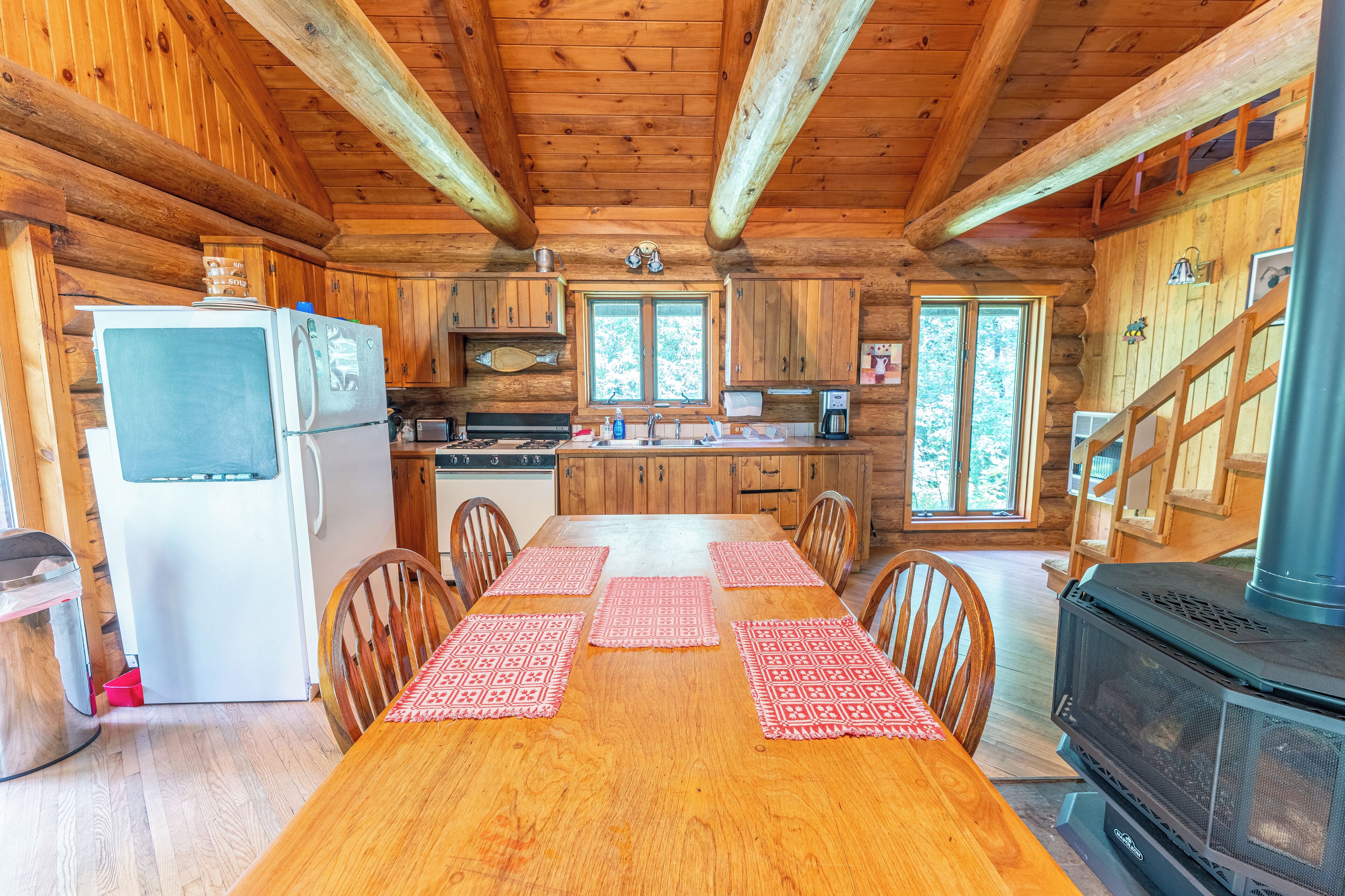 A wooden cabin interior with a dining table, chairs, a kitchen with a stove, and a refrigerator.