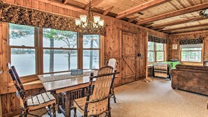 Ein rustikales Blockhaus-Interieur mit Esstisch, Holzstühlen, einer Chandelierleuchtre und großen Fenstern mit Seeblick.