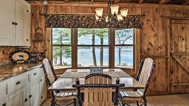 Ein rustikales Holzinterieur mit einem großen Esstisch, Stühlen und Blick auf einen See durch große Fenster.